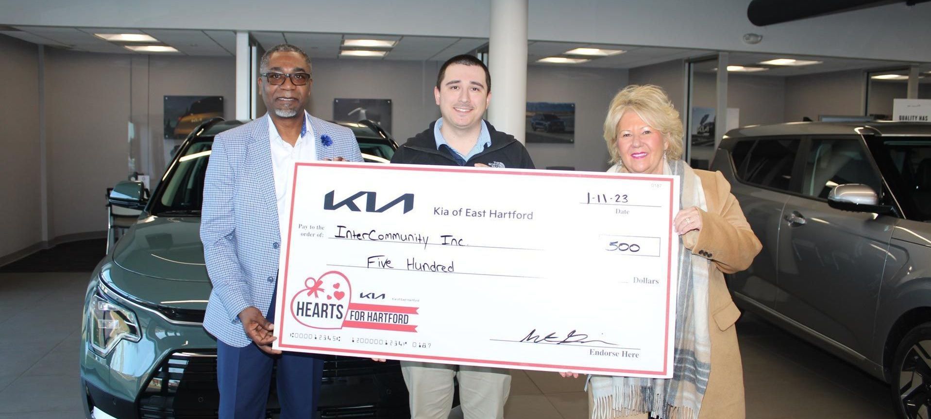 Three people in a dealership holding a large Kia ceremonial check for a donation.