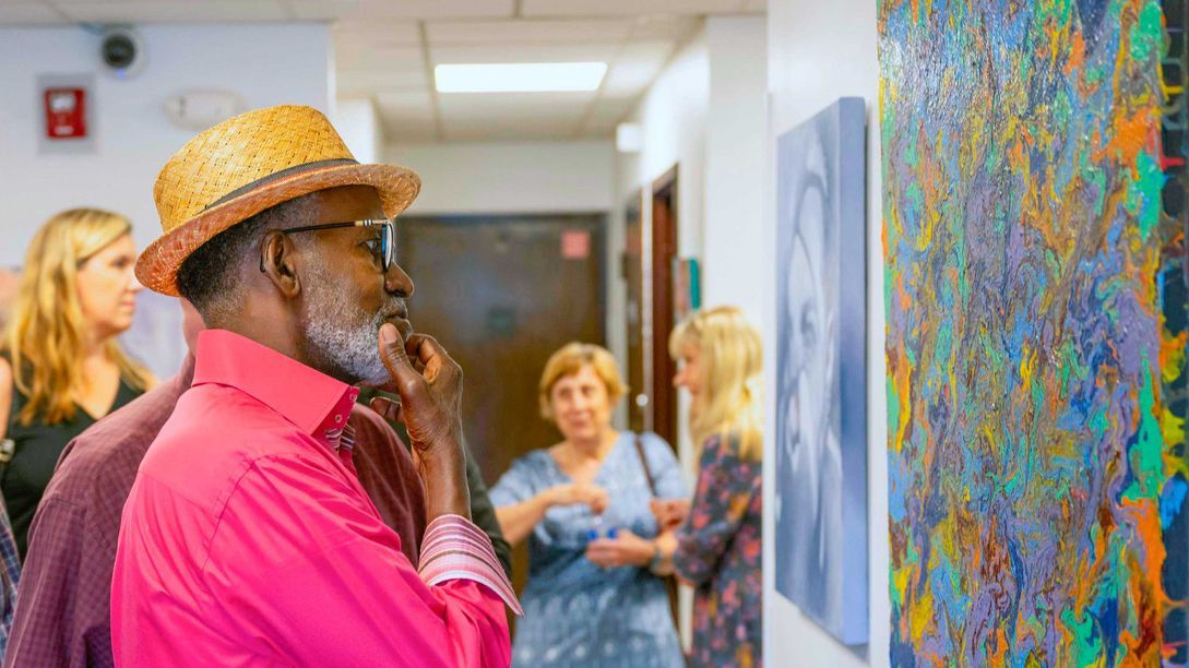 People look at paintings displayed on a wall in a brightly lit hallway at The Gallery.