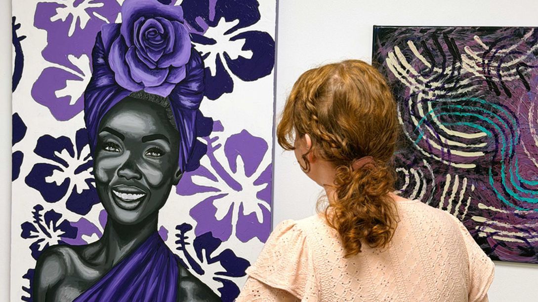 A person with auburn hair views a vibrant portrait of a smiling woman adorned with a large purple rose, beside abstract art.