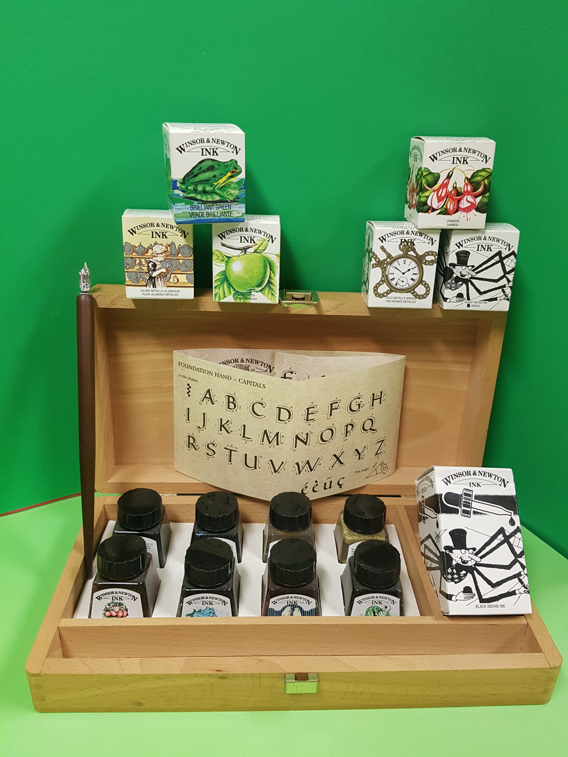Set di inchiostro in legno con flaconi, penna e scatole illustrative su sfondo verde.