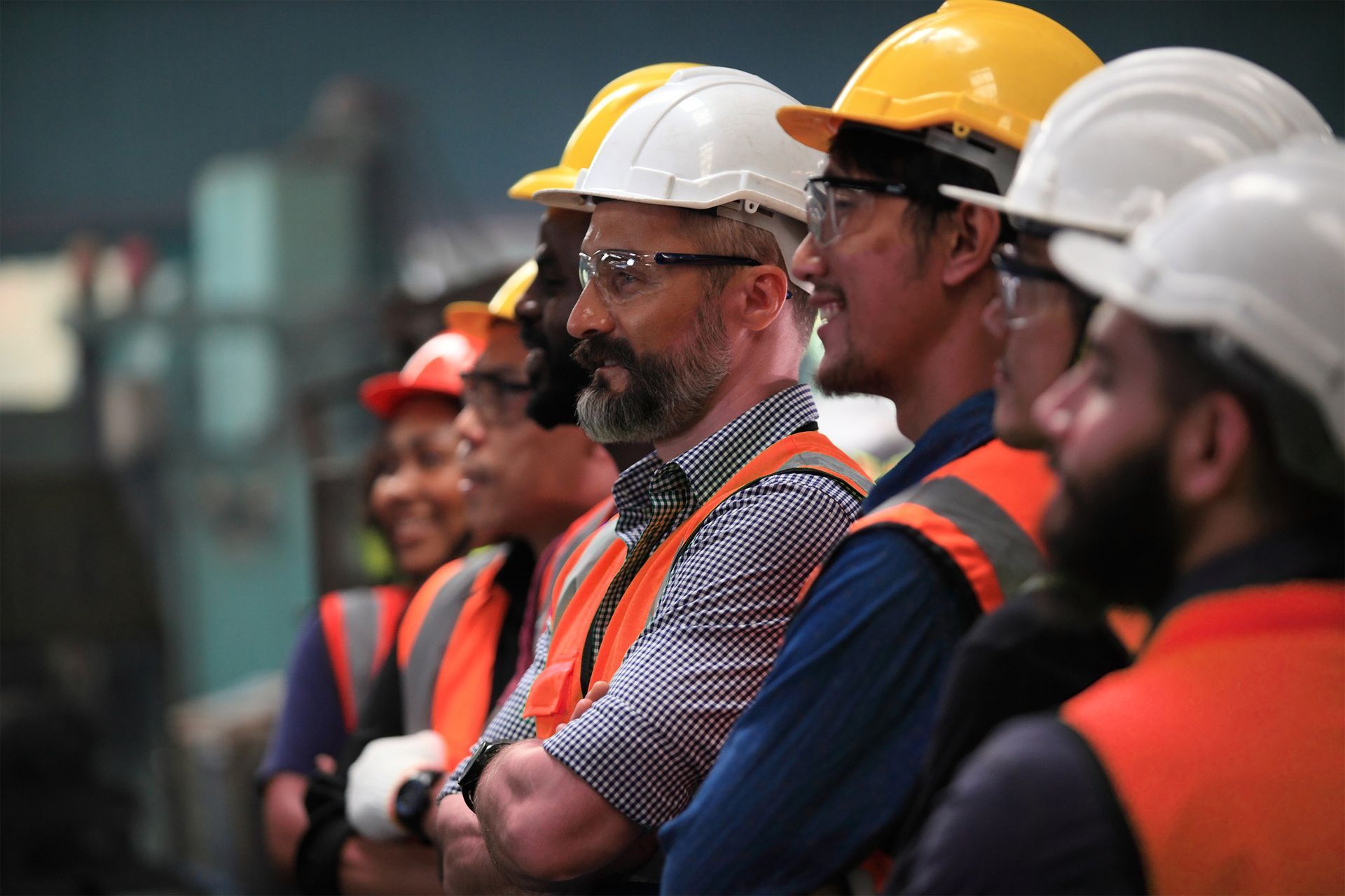 Des ouvriers, casqués et vêtus de gilets de sécurité, se tiennent debout dans une usine, le regard tourné vers l'horizon.