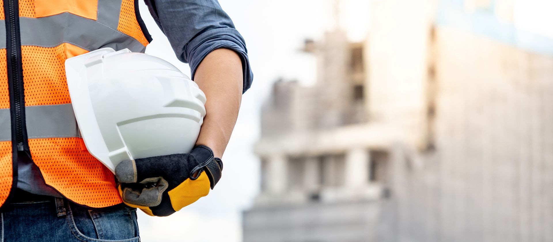 Un ouvrier du bâtiment, portant un casque blanc, un gilet réfléchissant et des gants, avec un bâtiment en arrière-plan.