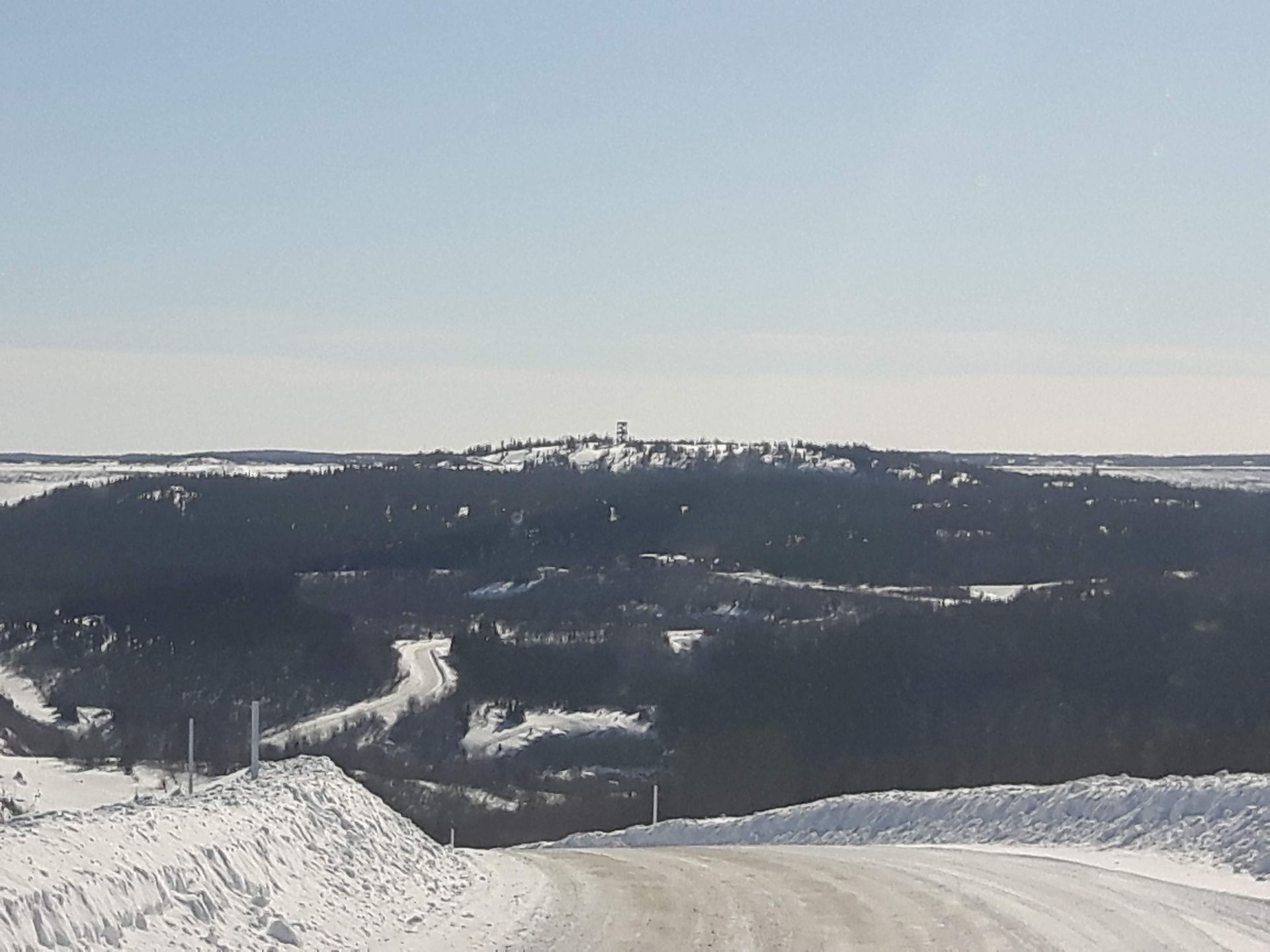 Paysage enneigé avec une route menant à une colline boisée surmontée d'une tour, sous un ciel d'un bleu éclatant.