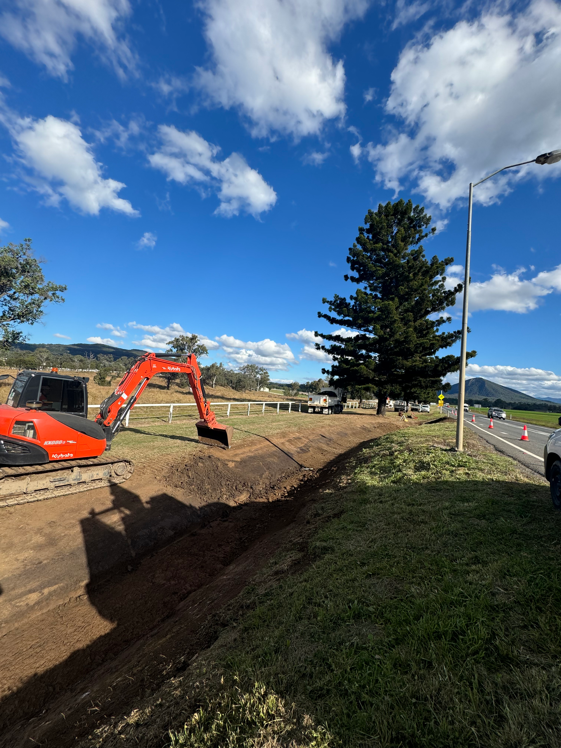 A red and black Spencer Civil & Earthworks excavator is working on a drainage ditch alongside a road.