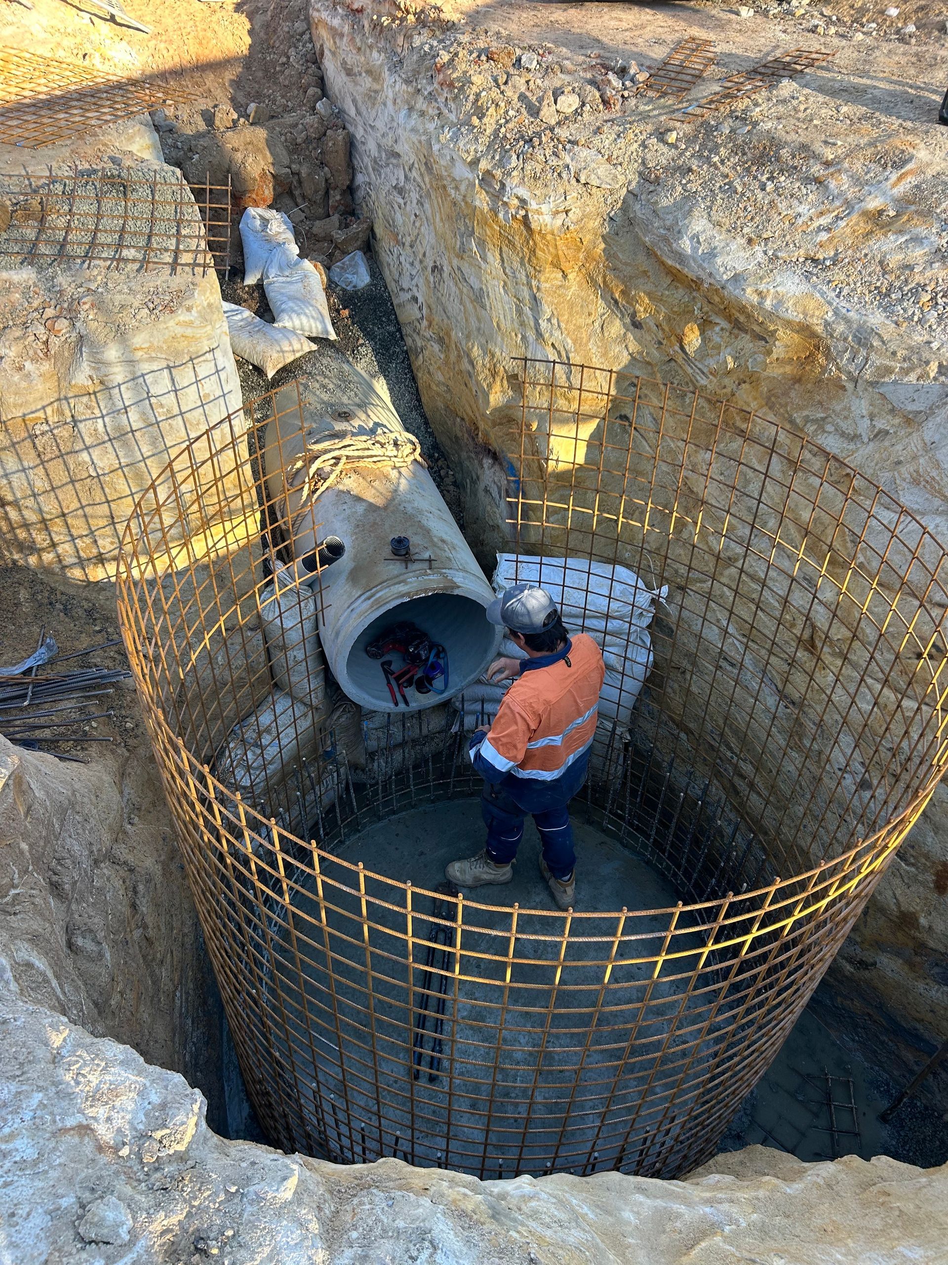 A man is standing in a hole with concrete reinforcing around the edge, inspecting a stormwater pipe.