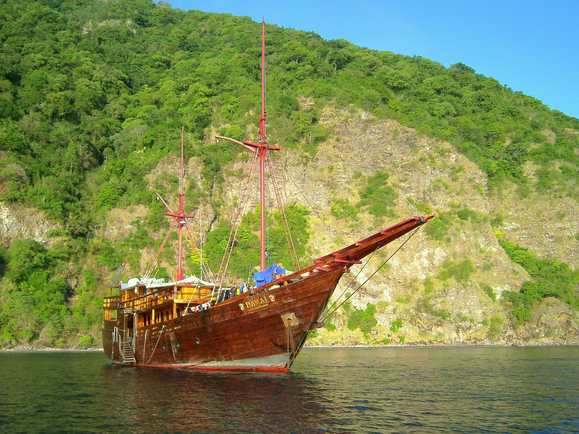 Old wooden ship floats on water near a green, mountainous shore.