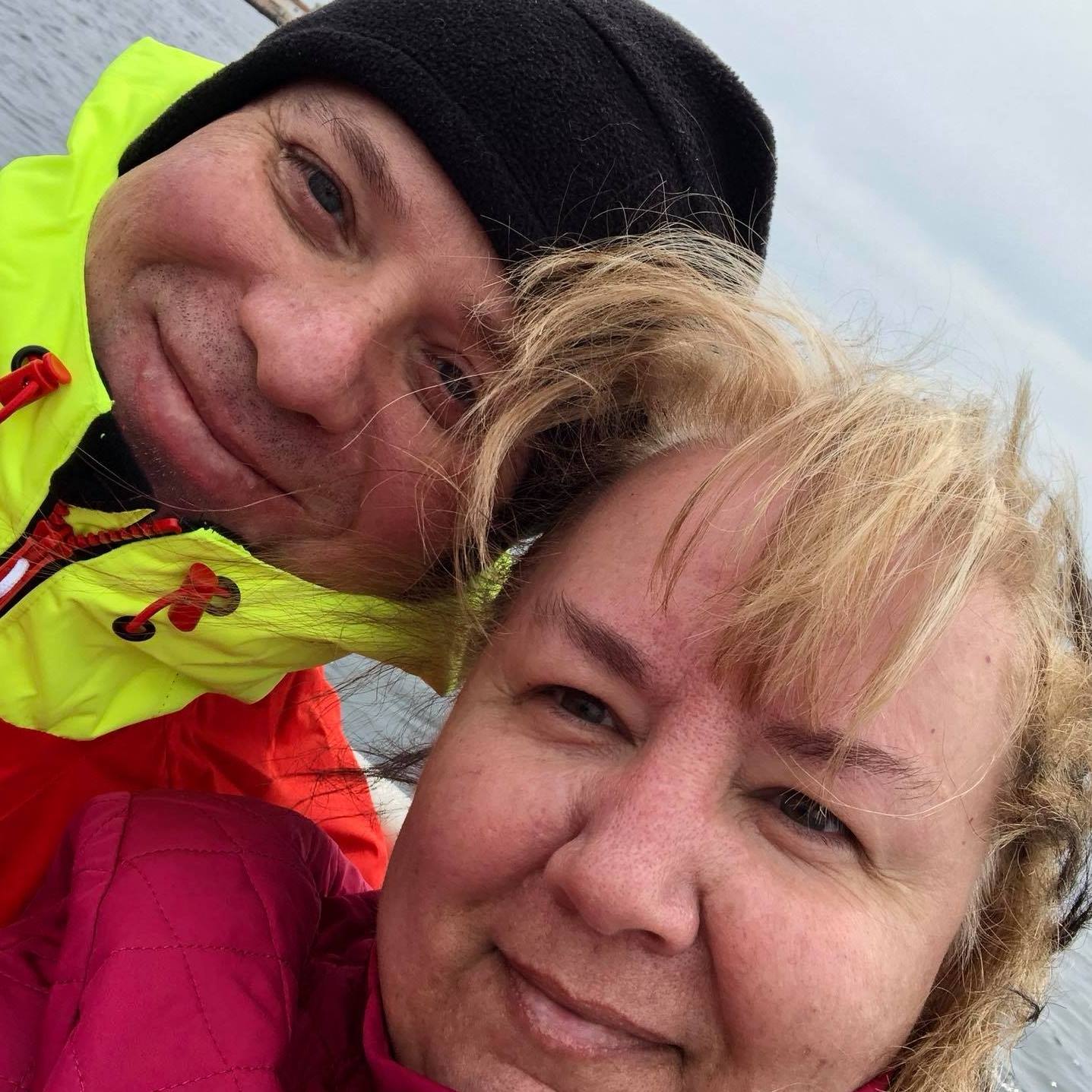 Couple in outdoor selfie; woman in pink, man in yellow jacket, both smiling, overcast.