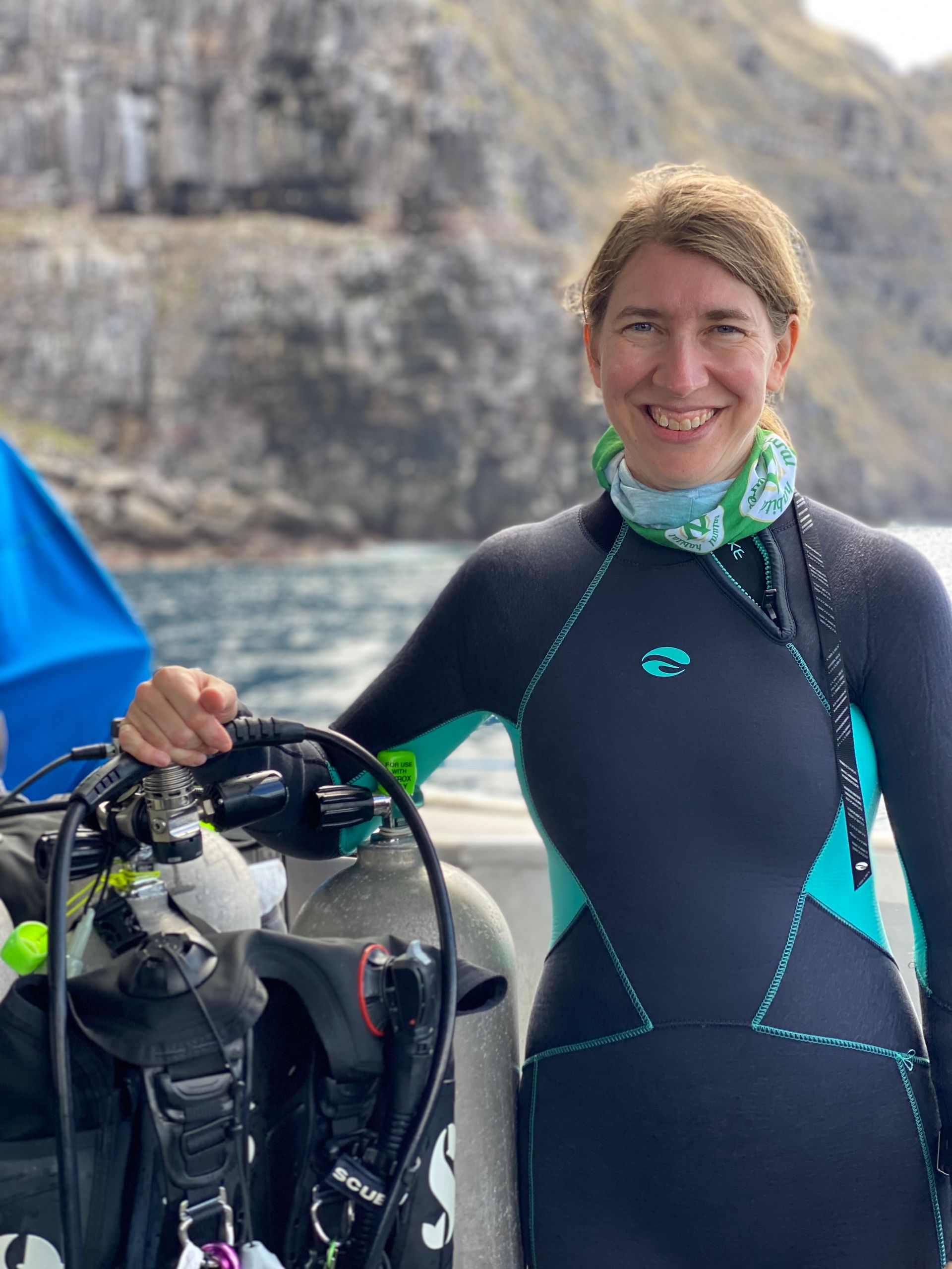 Woman in wetsuit with scuba gear on boat, smiling at camera near rocky shore.