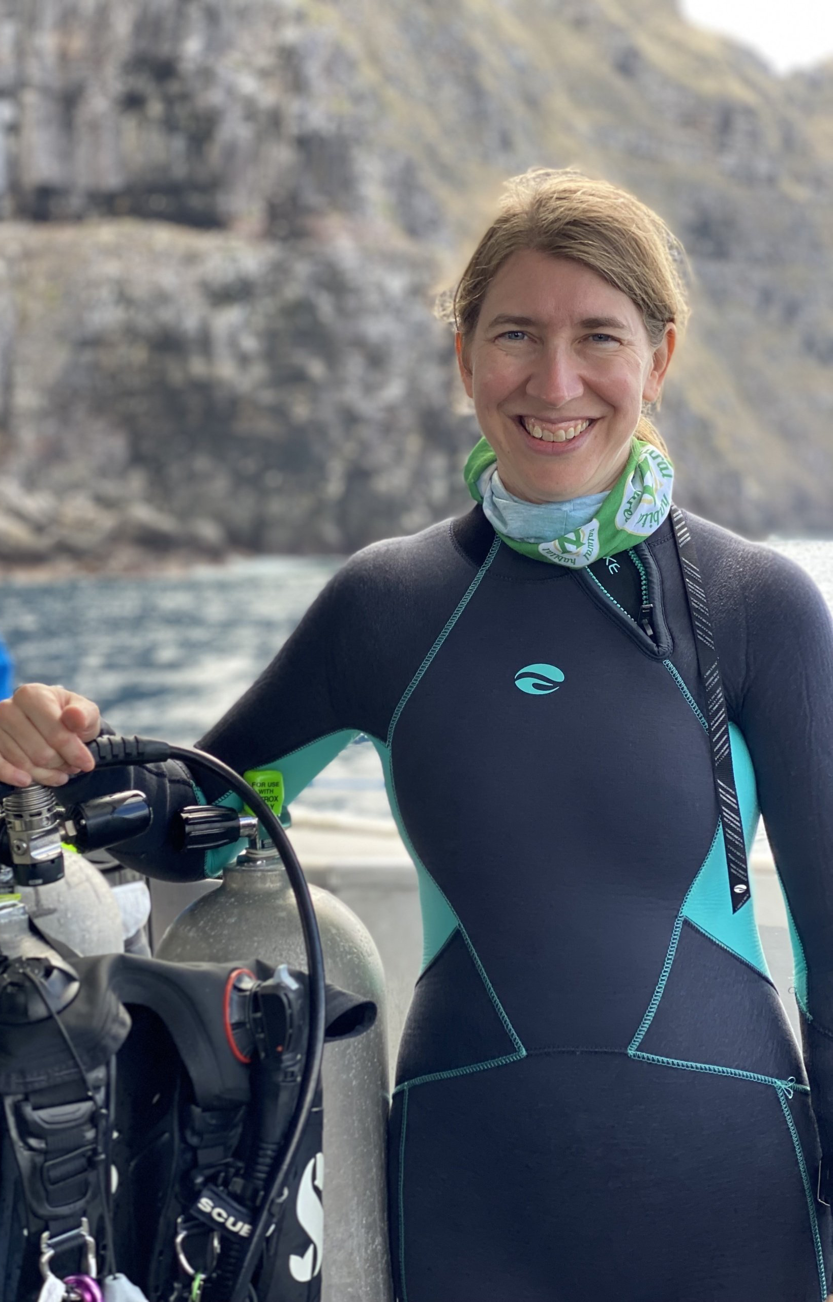 Woman in diving suit, holding scuba gear, smiling. Outdoors, ocean and rocky cliffs in background.