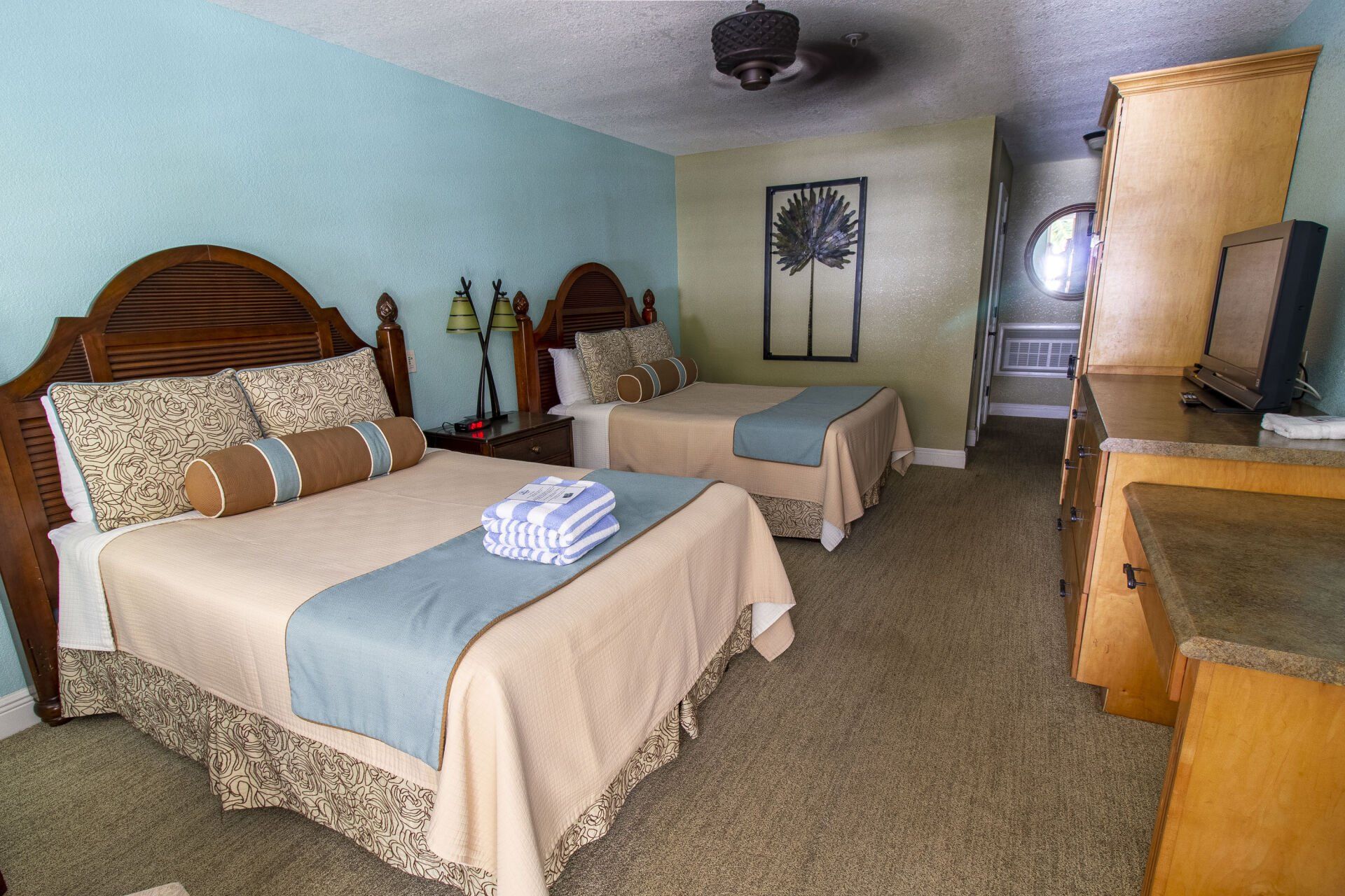 Hotel room with two beds, light blue walls, tan bedding, dresser, and decorative art.