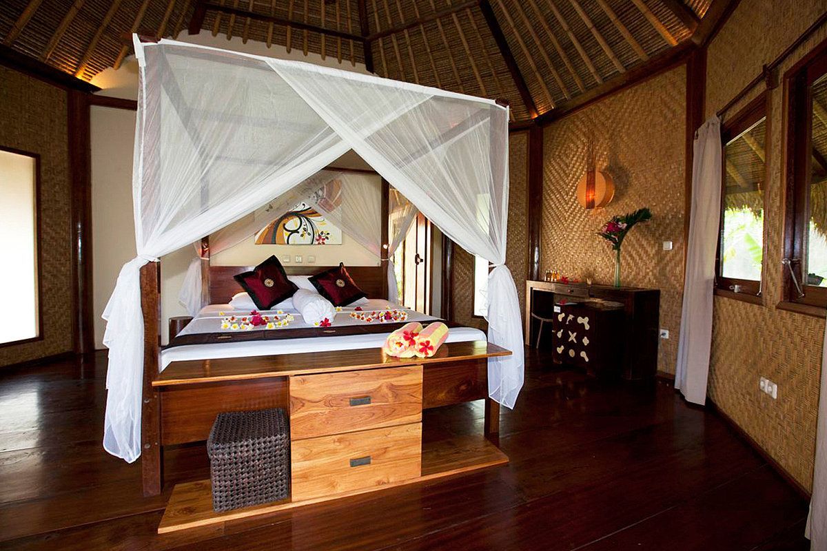Bedroom with wooden furniture, four-poster bed with mosquito net, straw roof and woven walls.