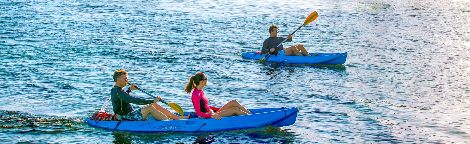 Two kayaks on water, each with a person paddling. One kayak has two people. Blue water, sunny day.