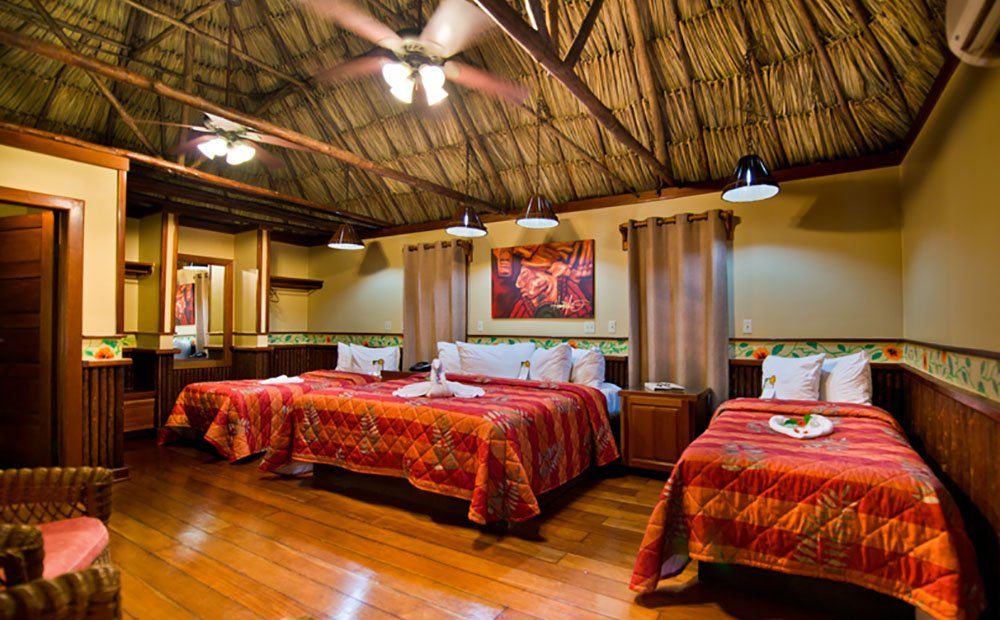 Spacious hotel room with three beds, wooden floor, and thatched ceiling.