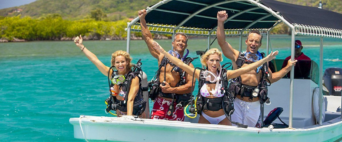 Four people in scuba gear celebrate on a boat in turquoise water.