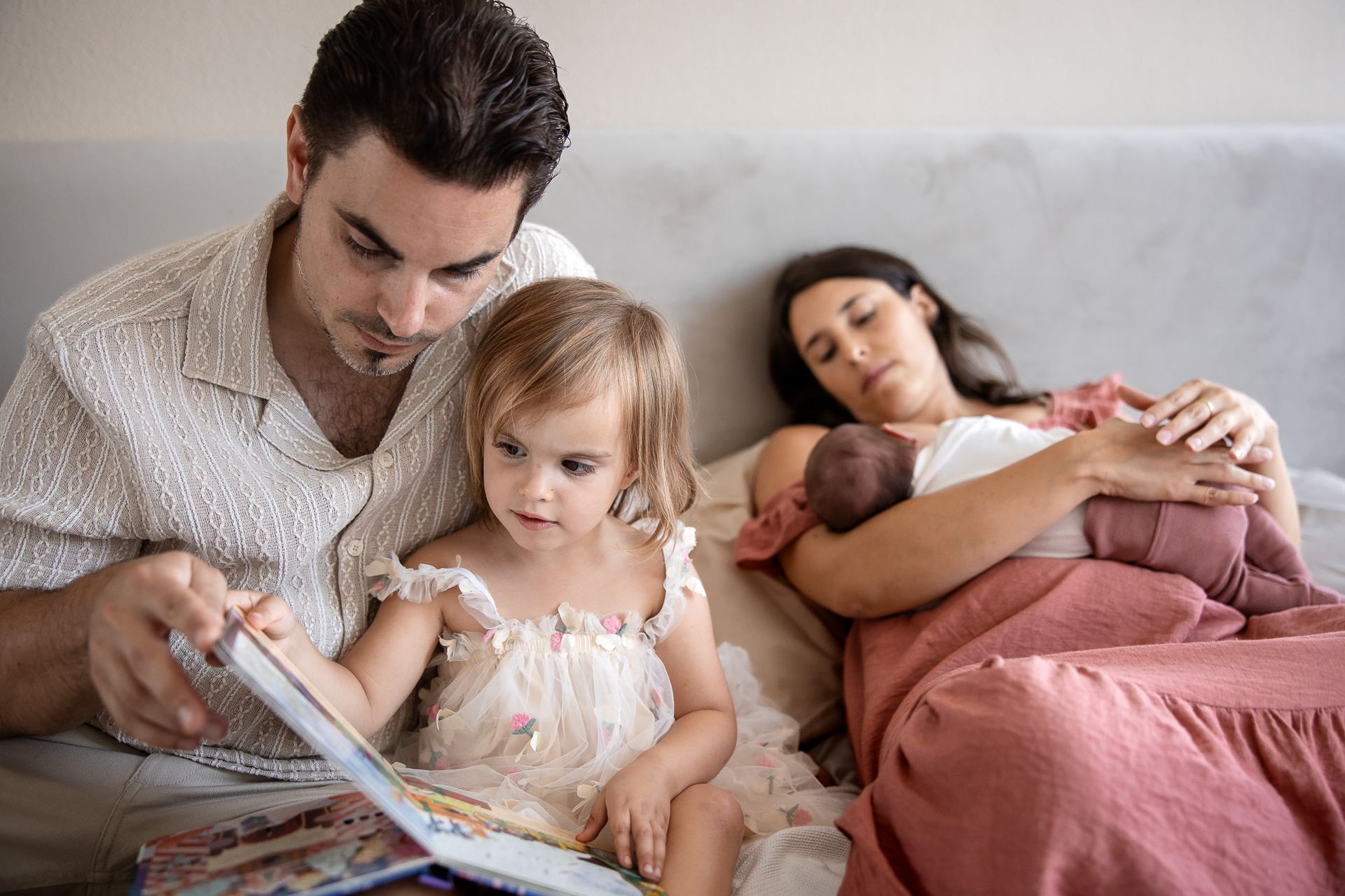 Vader leest voor aan peuter terwijl moeder met baby op bed ligt