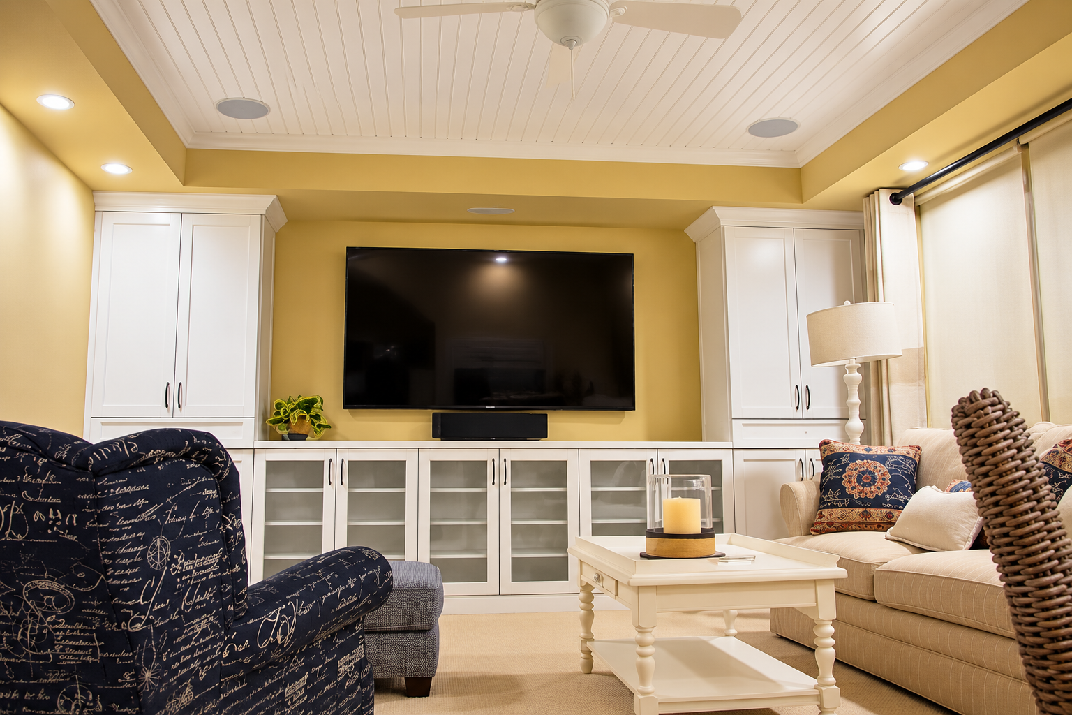 Cozy living room with a large TV, white built-ins, cream sofa, patterned chair, and warm recessed lighting