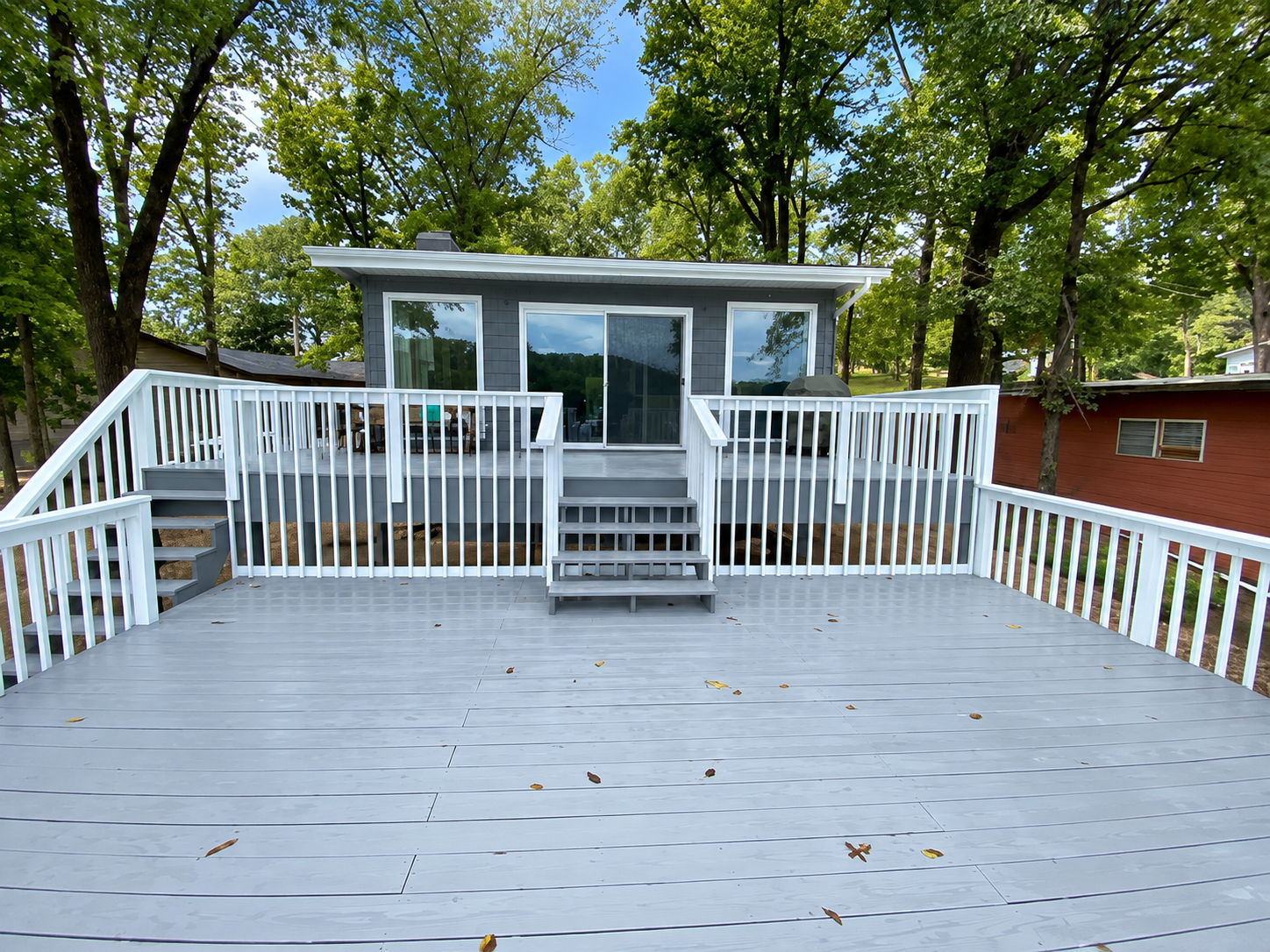Gray wooden deck leading to a small white house with railing and trees in the background