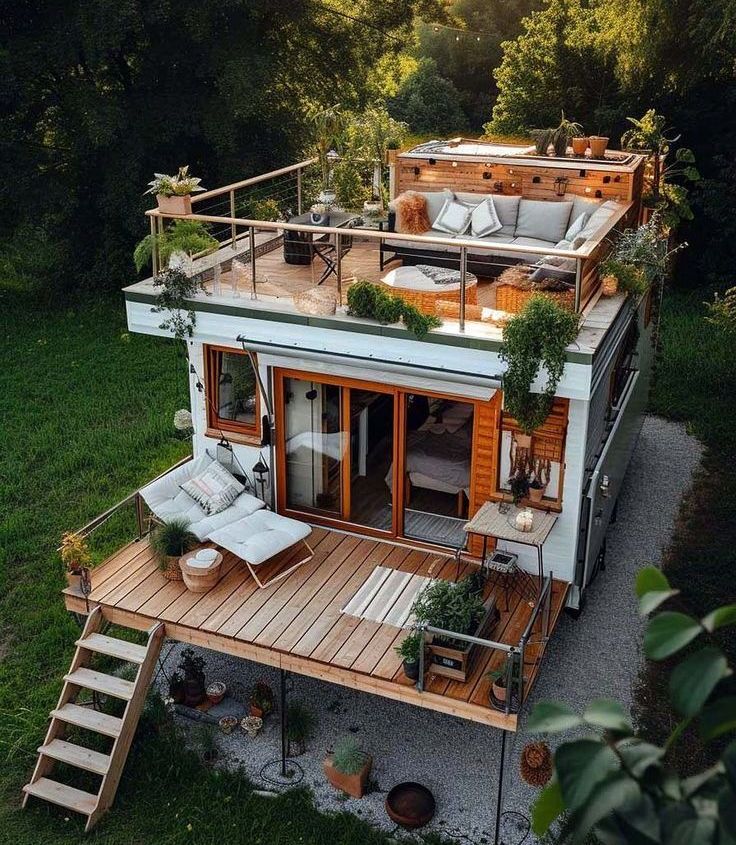 Tiny white house with rooftop deck, surrounded by greenery. Wooden decks, outdoor furniture, and potted plants.
