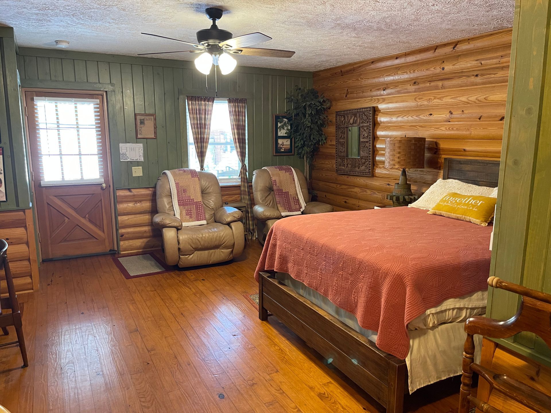 A rustic bedroom with wooden walls, a bed, two recliners, and a door leading outside.