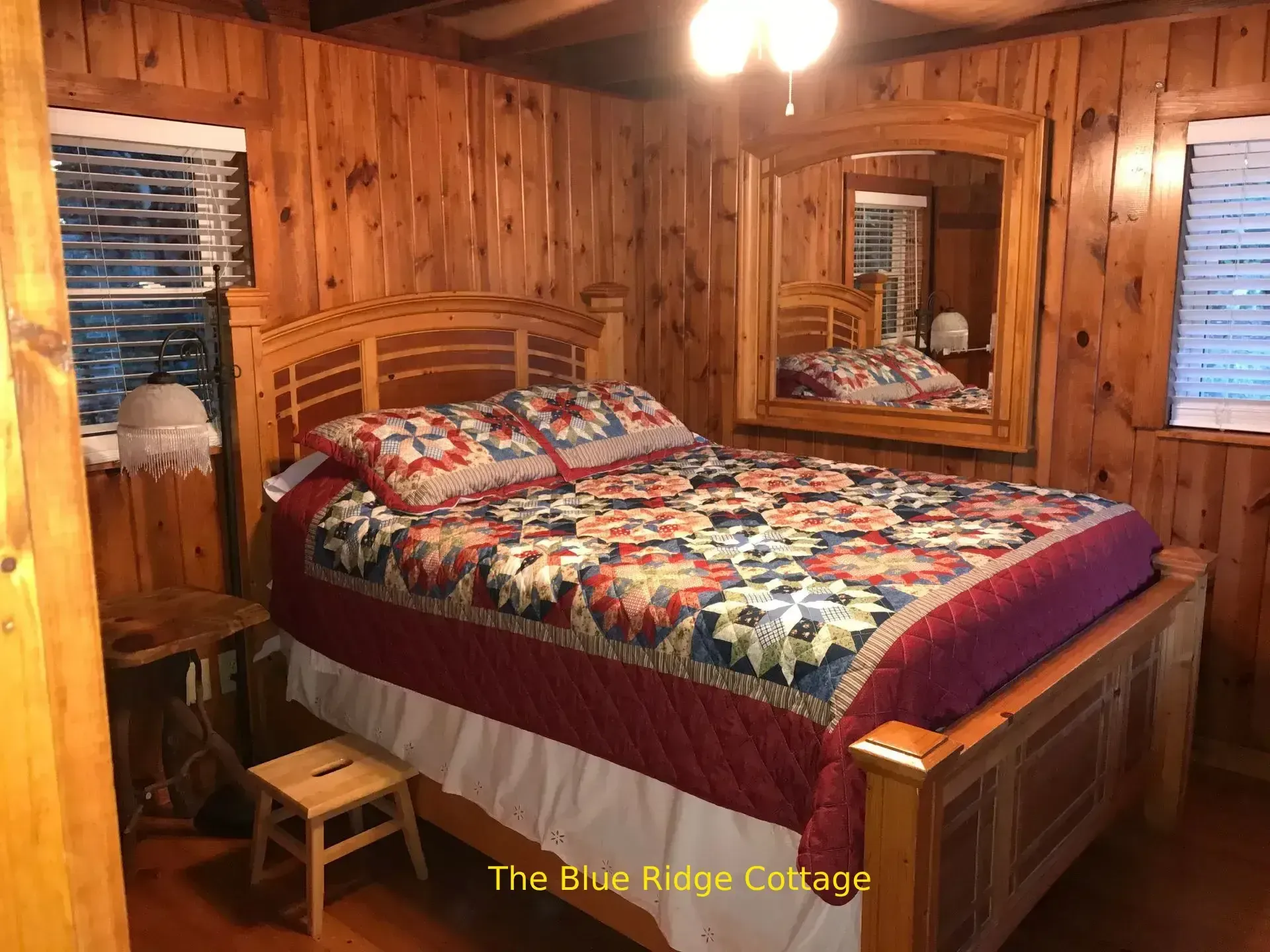 Wooden-paneled bedroom with floral quilt, bed, mirror, window, and small stool.