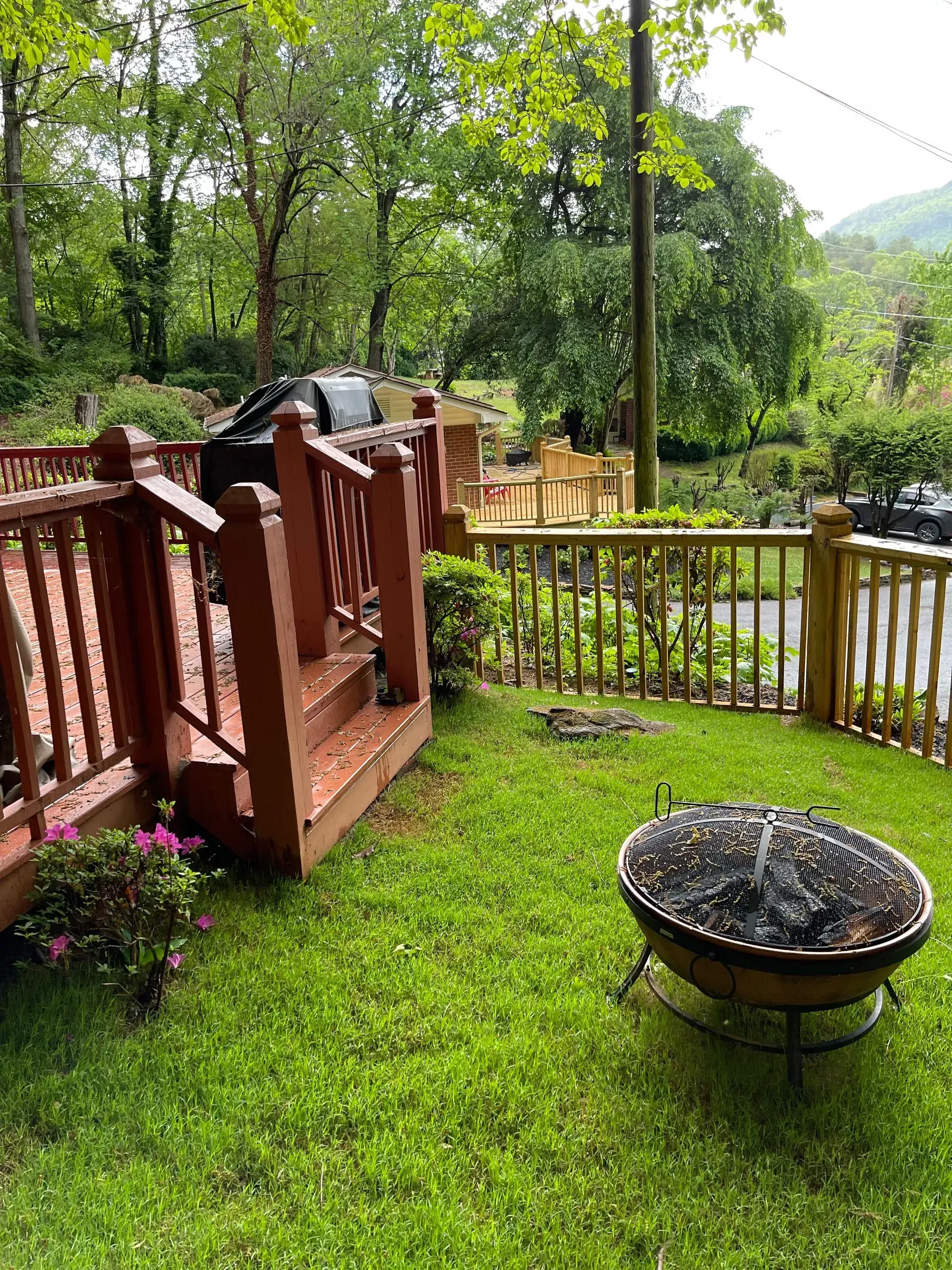 A deck with red railing and steps, grass, a fire pit, and a wooden fence. Green trees in the background.