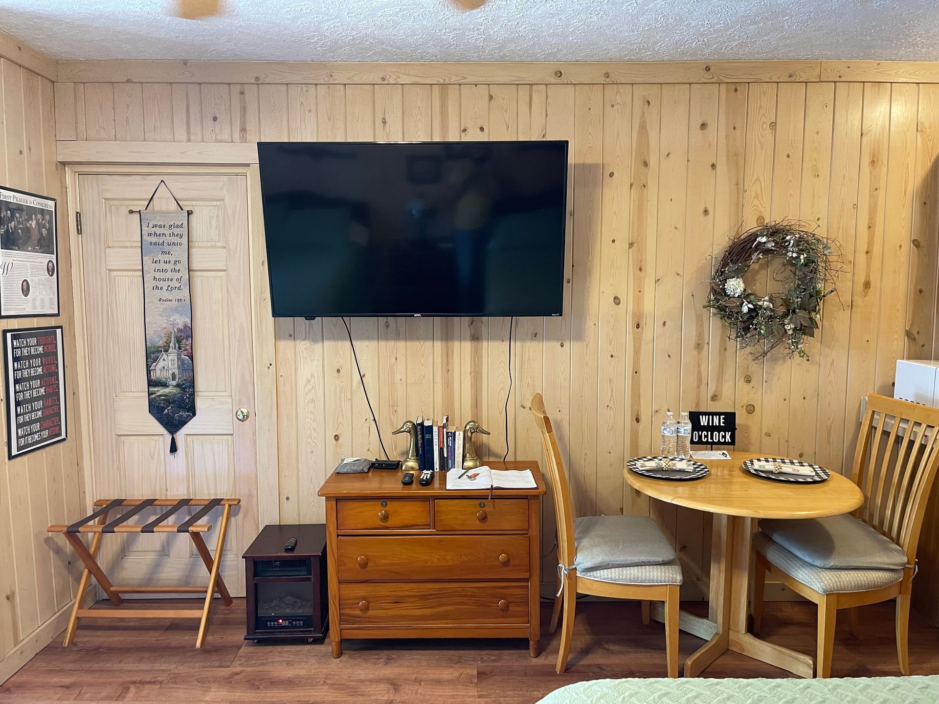 A small room with wood-paneled walls features a TV, dresser, table with chairs, and a door.