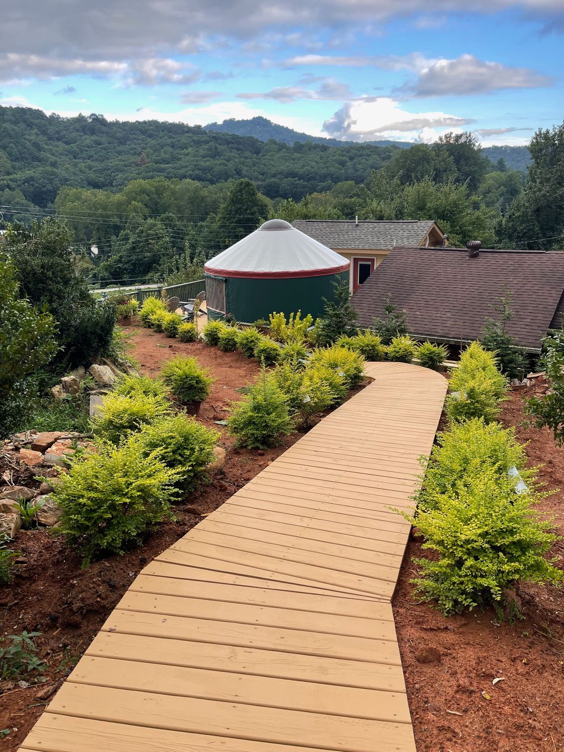 Wooden walkway leads to a yurt and building surrounded by greenery, with a mountain backdrop.