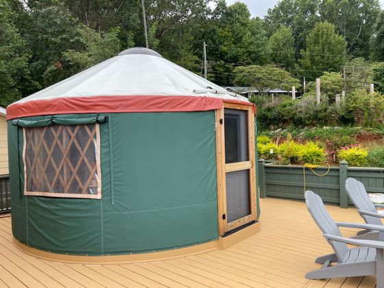A green yurt with a wooden door and a red-trimmed roof, set on a wooden deck.