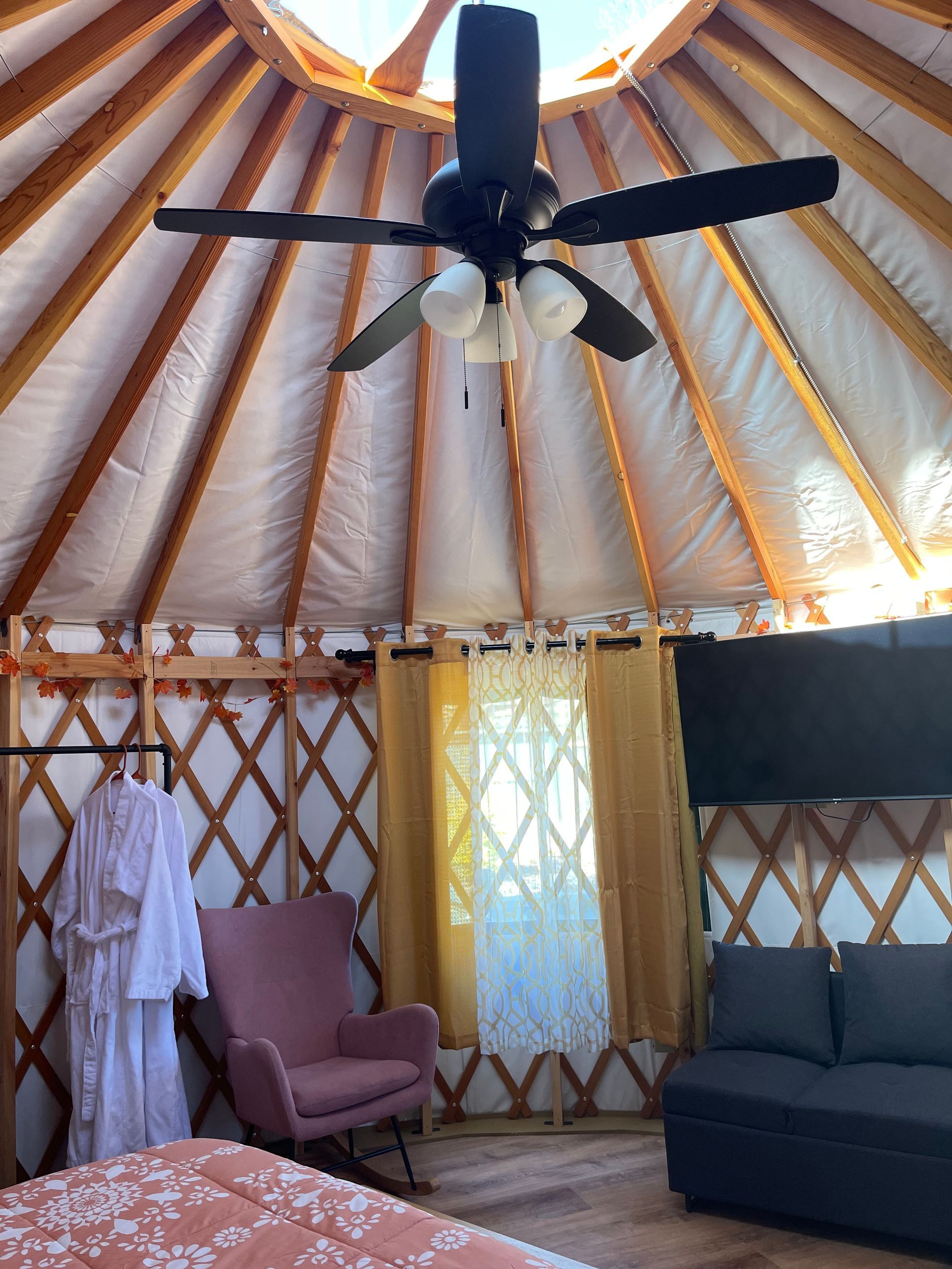 Inside of a yurt. A ceiling fan, skylight, and TV. Wooden lattice walls with pink chairs.