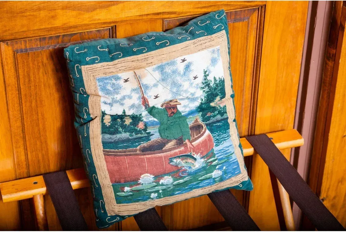 Pillow on luggage rack: man fishing from boat, green jacket, blue water, forest backdrop.