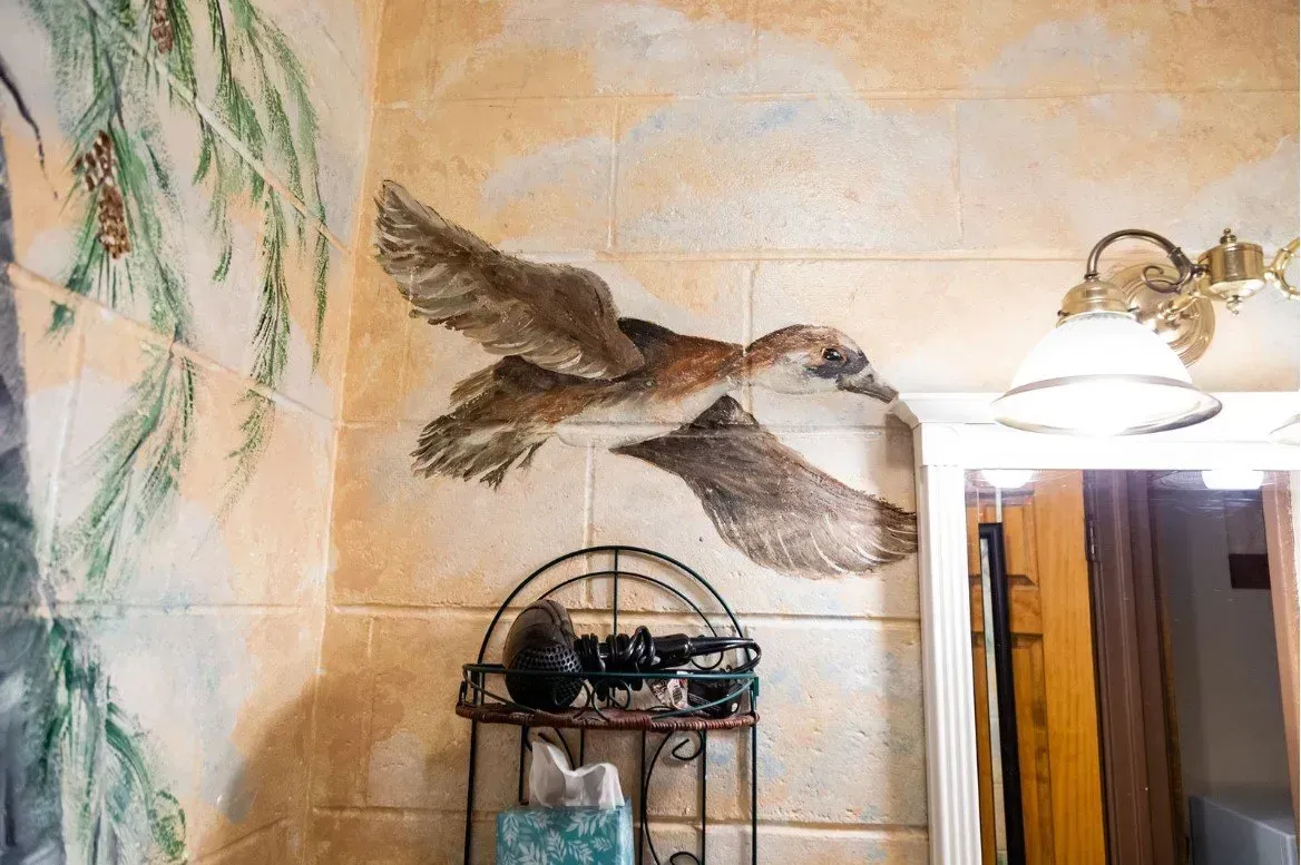 Mural of a duck in flight, painted on a textured wall. Brown and beige tones, near a small shelf.