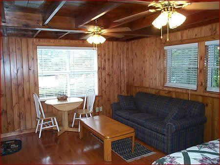 Cozy cabin interior: wood paneling, sofa, round table with chairs, coffee table, ceiling fans, windows with blinds.