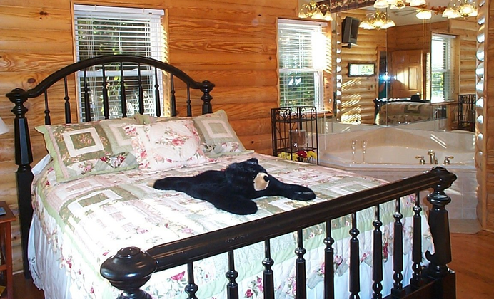 Cozy bedroom with a black bed frame, floral quilt, and a jacuzzi visible through a doorway. A plush bear lies on the bed.