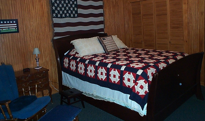 Bedroom with a large bed, American flag headboard, patriotic quilt, blue chair, and straw wall.