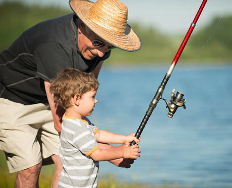 Grandfather teaching a young boy to fish by a lake; both are smiling.