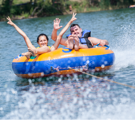 Two people on an orange and blue inflatable tube being towed on a lake, smiling and waving.