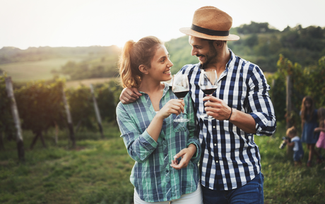 Couple in a vineyard toasting wine, smiling at each other. Man has a hat and striped shirt. Woman has a ponytail.