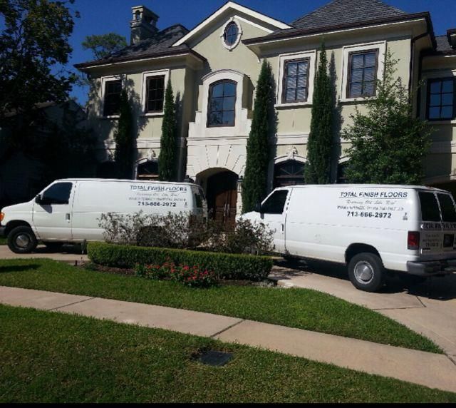 Two white vans parked in front of a large, tan house with tall cypress trees.