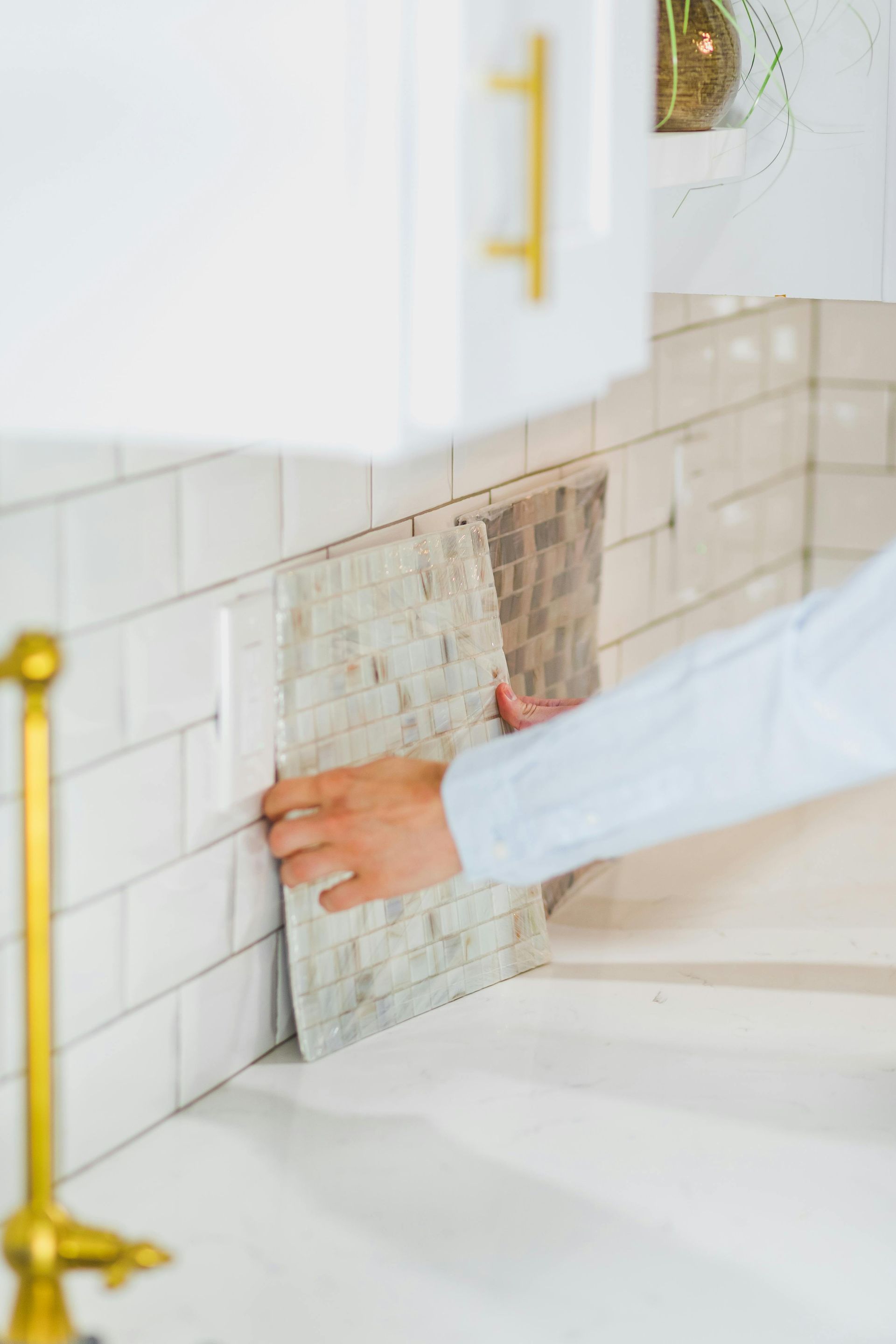 Person installing mosaic tile backsplash in white kitchen, holding tile against white subway tile.