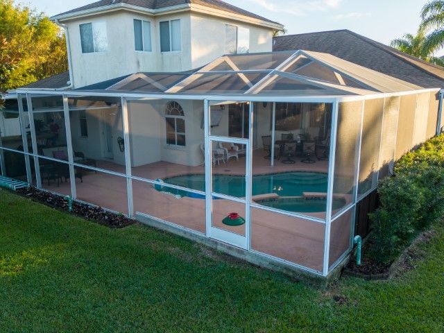 A white-framed pool cage enclosure attached to a two-story house with a swimming pool inside, viewed from the lawn.