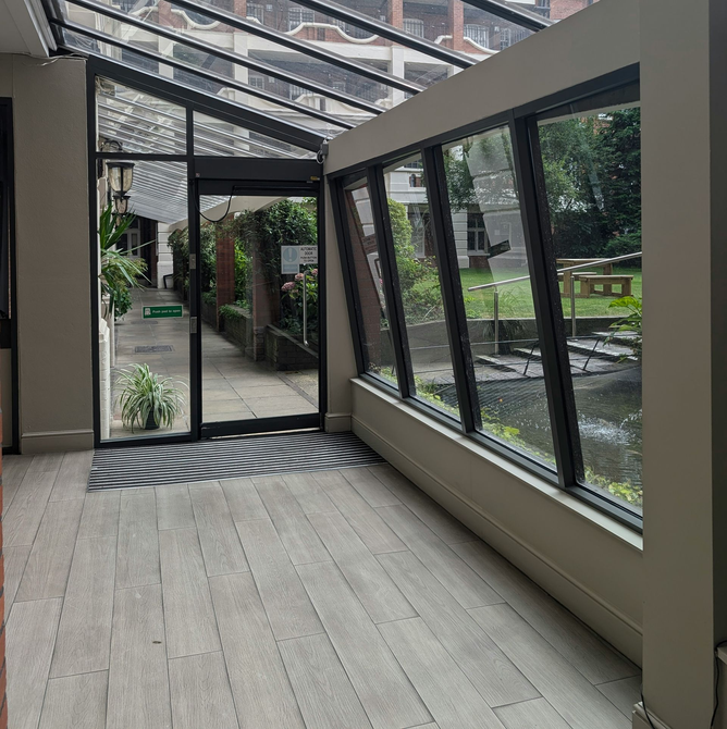 A glass-walled walkway with a wood-look floor, leading to a doorway that opens onto a garden courtyard.