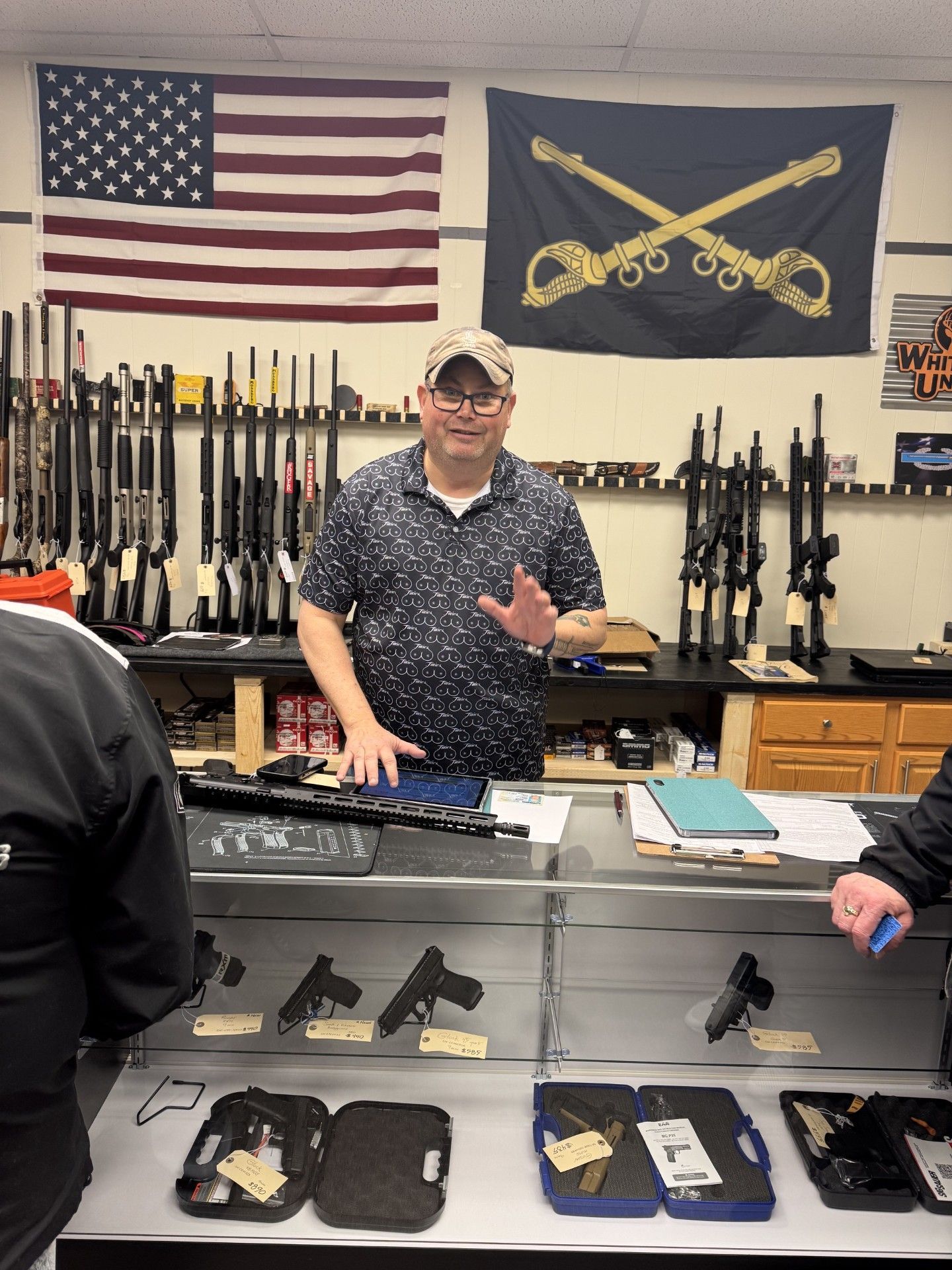 A customer stands behind the counter inside a firearm retail store, with rifles displayed on wall racks behind him, handguns showcased in a glass display case, and an American flag alongside a crossed-sabers flag hanging on the wall.