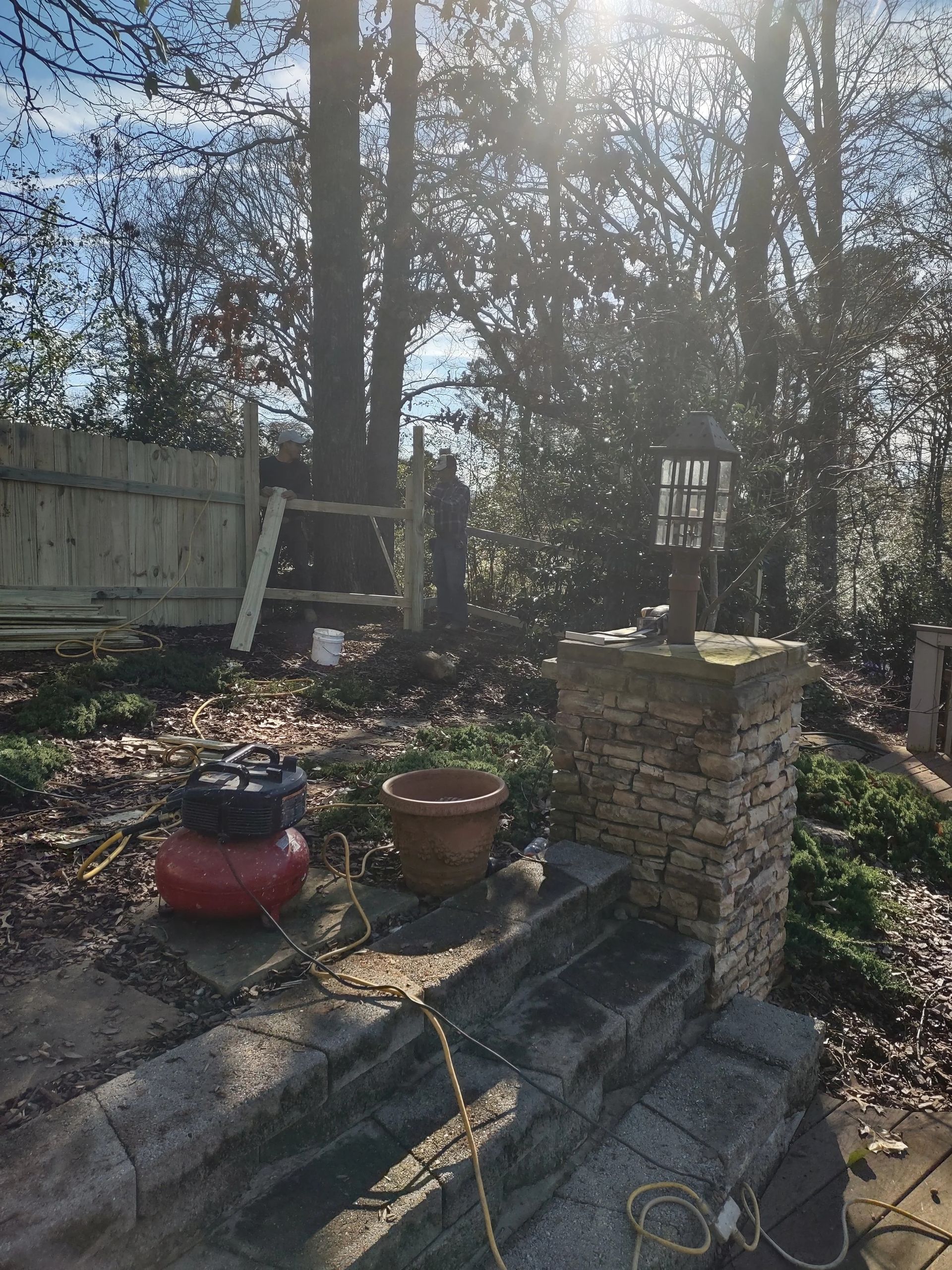 Backyard scene: stone steps, brick pillar with lamp, air compressor, tree, wooden fence, ladder.