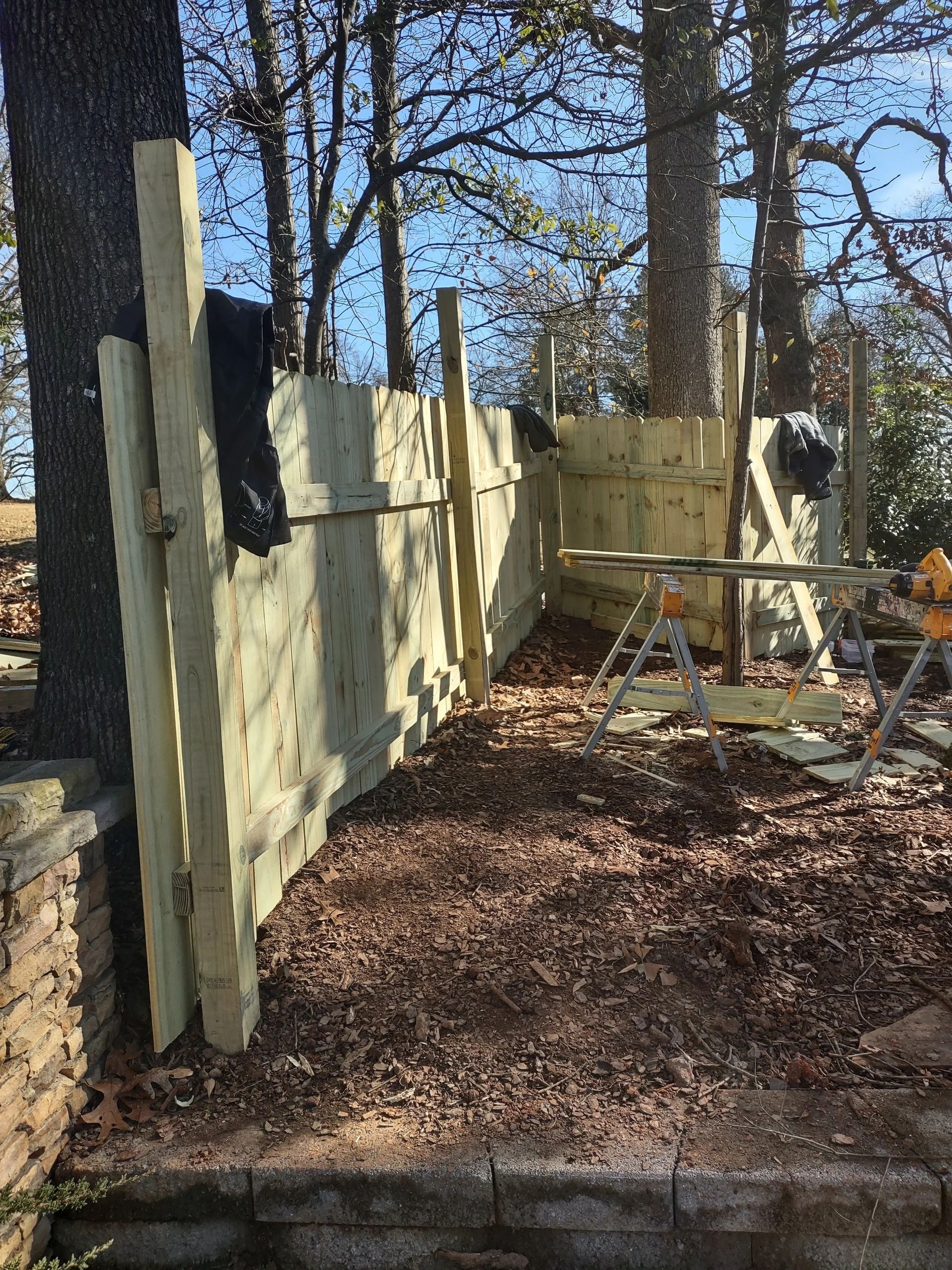 Wooden fence construction beside trees, with sawdust ground cover, and a partial view of construction equipment.