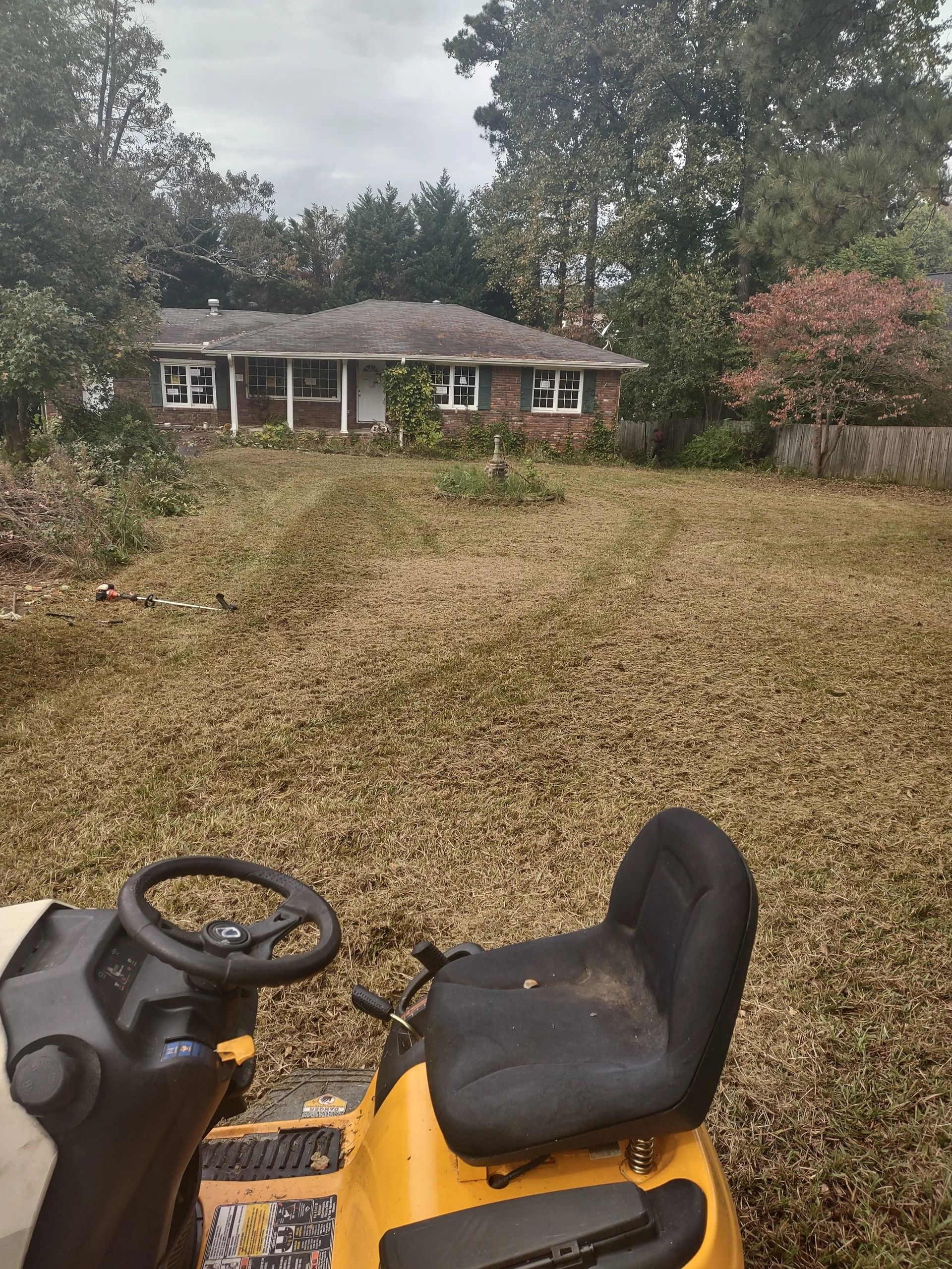 Yellow riding lawnmower in a yard, cutting dry grass in front of a brick house. Overcast sky.