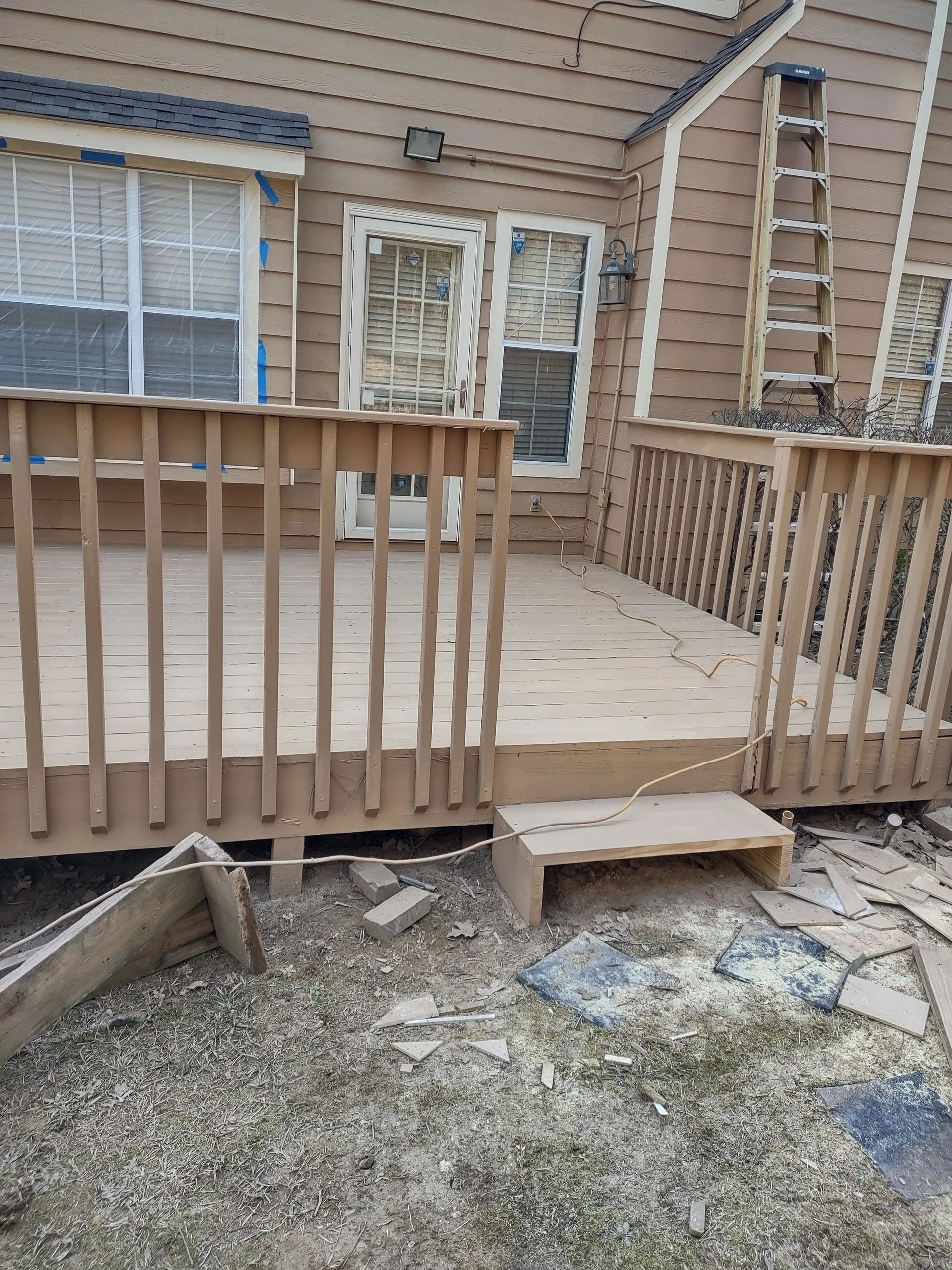 Wooden deck with railing and steps, leading to French doors. Brown siding and ladder are visible.