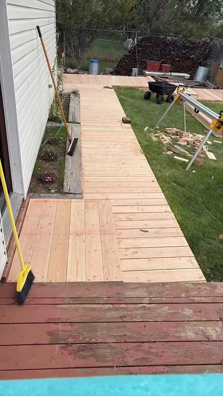 Backyard wooden walkway under construction beside a white house, leading to a grassy yard with tools and sawhorses.