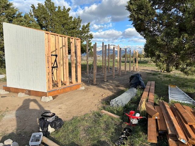 Partially built wooden shed frame in a rural yard with lumber and tools in the foreground