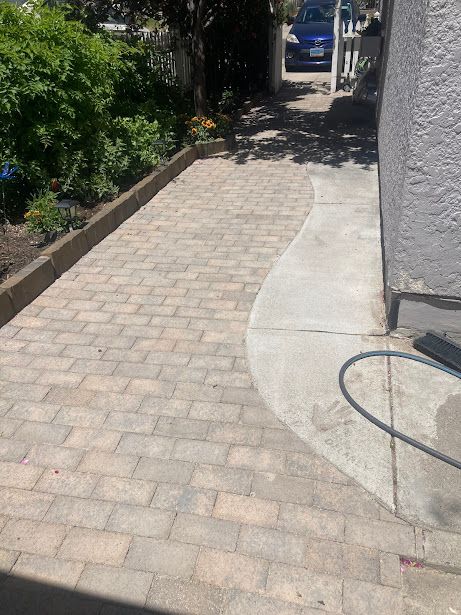 Sunlit brick walkway beside a stucco wall and garden, with a hose on the right and trees ahead