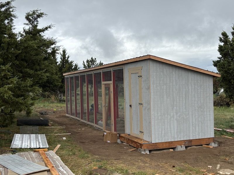 Small white shed-like structure on a dirt lot under cloudy skies, with trees nearby.